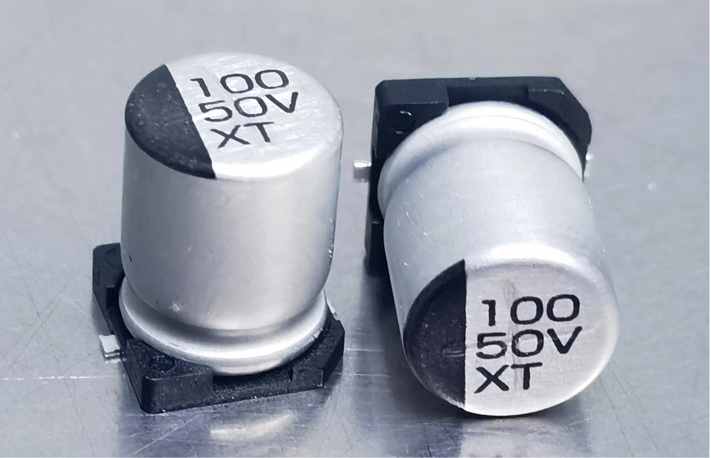 Close-up of two SMD electrolytic capacitors (Surface Mount Device) with black stripe and silver top, clearly marked 100 μF 50V XT rating for testing key points.